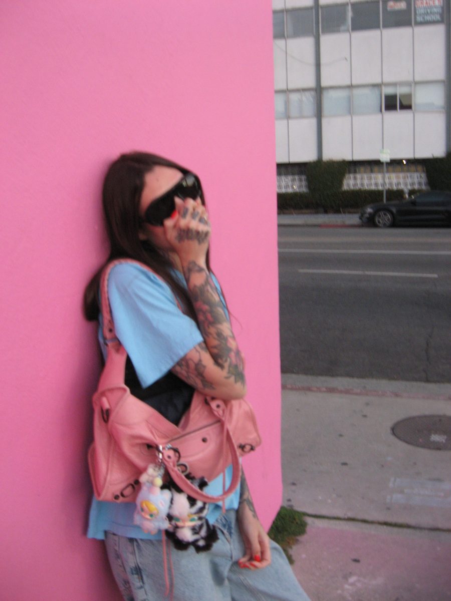 Sasha in sunglasses and a baggy tee against the pink wall